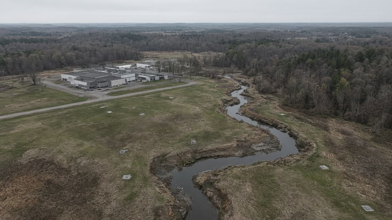 Aerial view of an industrial facility, grass well pads, and a creek winding through the floodplain.